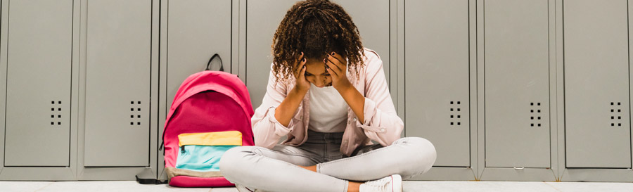 A young black girl sitting alone in a school hallways with her head in her hands.