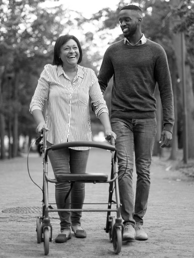Photo of disabled woman in a walker. A man is next to her helping out. They are both enjoying a nice day at the park.