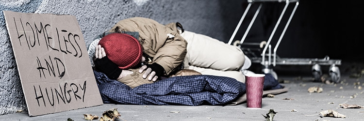 Photo of a homeless man sleeping in a alley with a sign next to him with hand written words saying Homeless and Hungry.