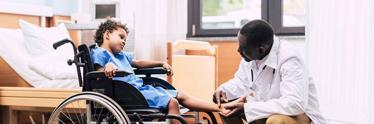 Photo of a young child in a wheelchair with a doctor kneeling down looking at his leg.