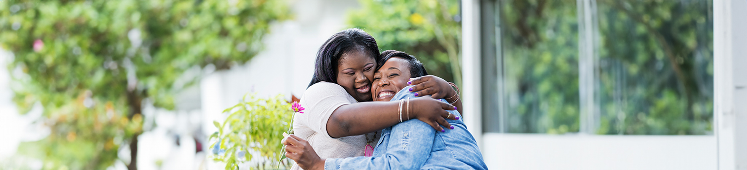 A black mother holding a pink flower with her teenage daughter who has down syndrome. They are outdoors, in the back yard of their home, smiling and hugging at the camera.