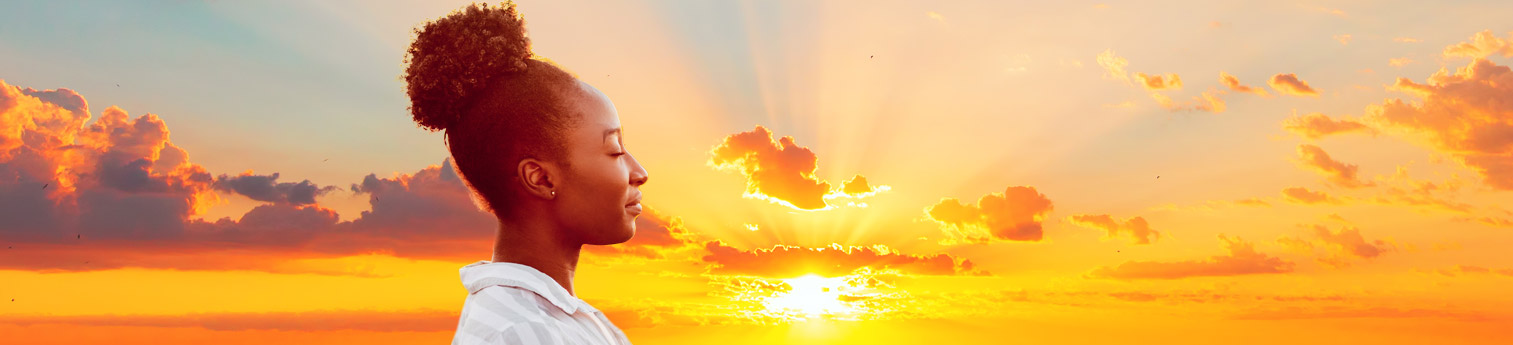 a young black woman smiling with her eyes closed. She is happy while she is outside enjoying a sunset.