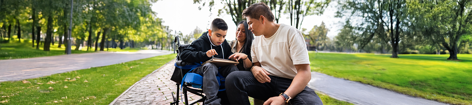 A Latino child in a wheelchair, with his mother and father reading a book in a park.