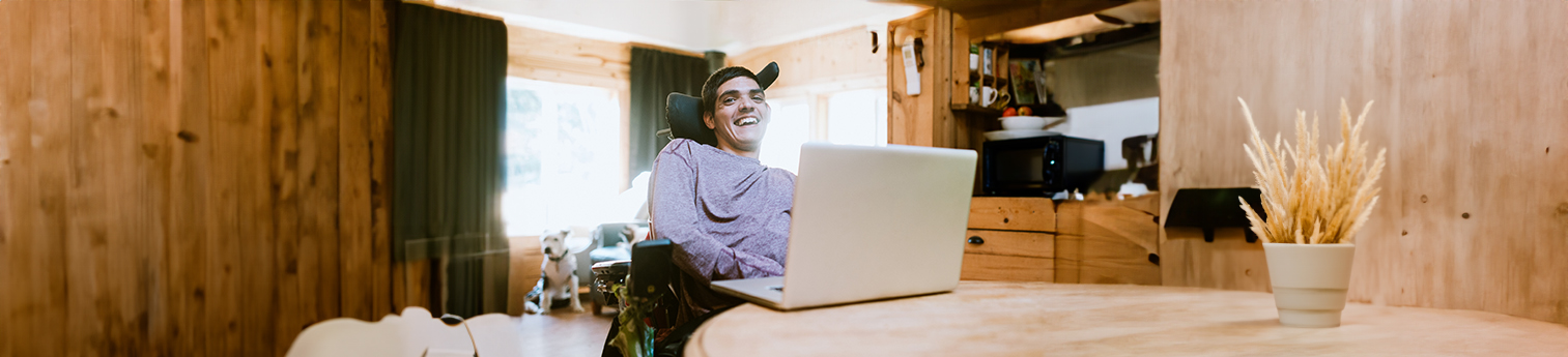 A cheerful young adult man with cerebral palsy on his laptop, with a pit bull in the background