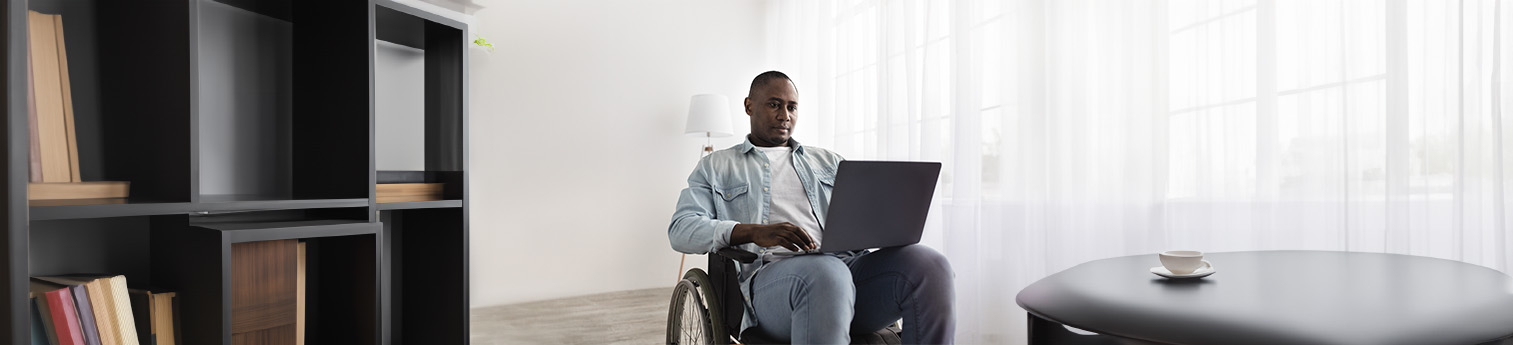 A black male in a wheelchair with his laptop on his lap, in a living room with a bookshelf and a table with a cup of coffee