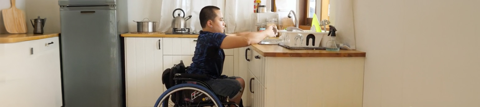 An Asian man in a wheelchair in his kitchen washing dishes.