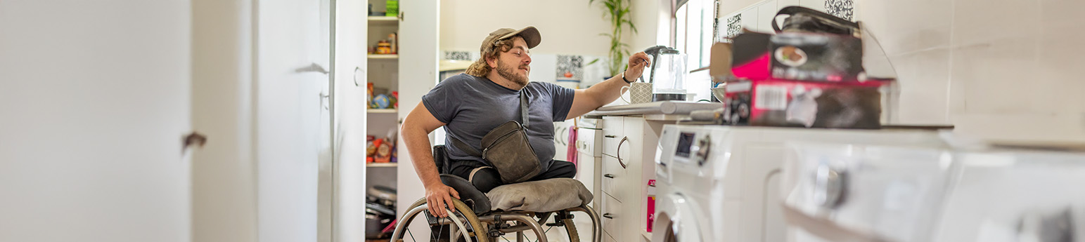A white man in a wheelchair with blonde hair wearing a cap, he has both of his legs amputated and is in kitchen reaching over for his mug.