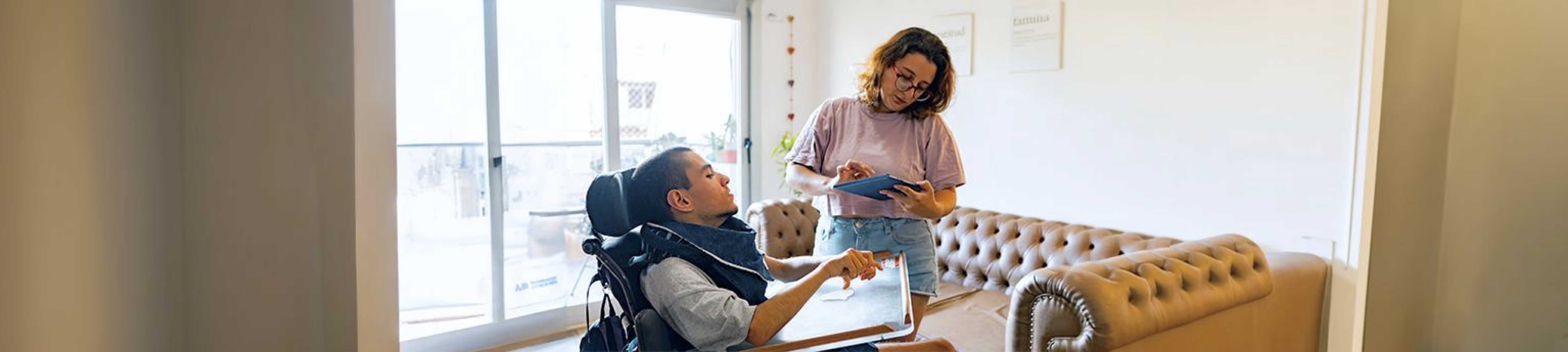A photo of a latina sister and a disabled latino brother in wheelchair in their apartment. The sister is on her electronic tablet with a stressed expression.