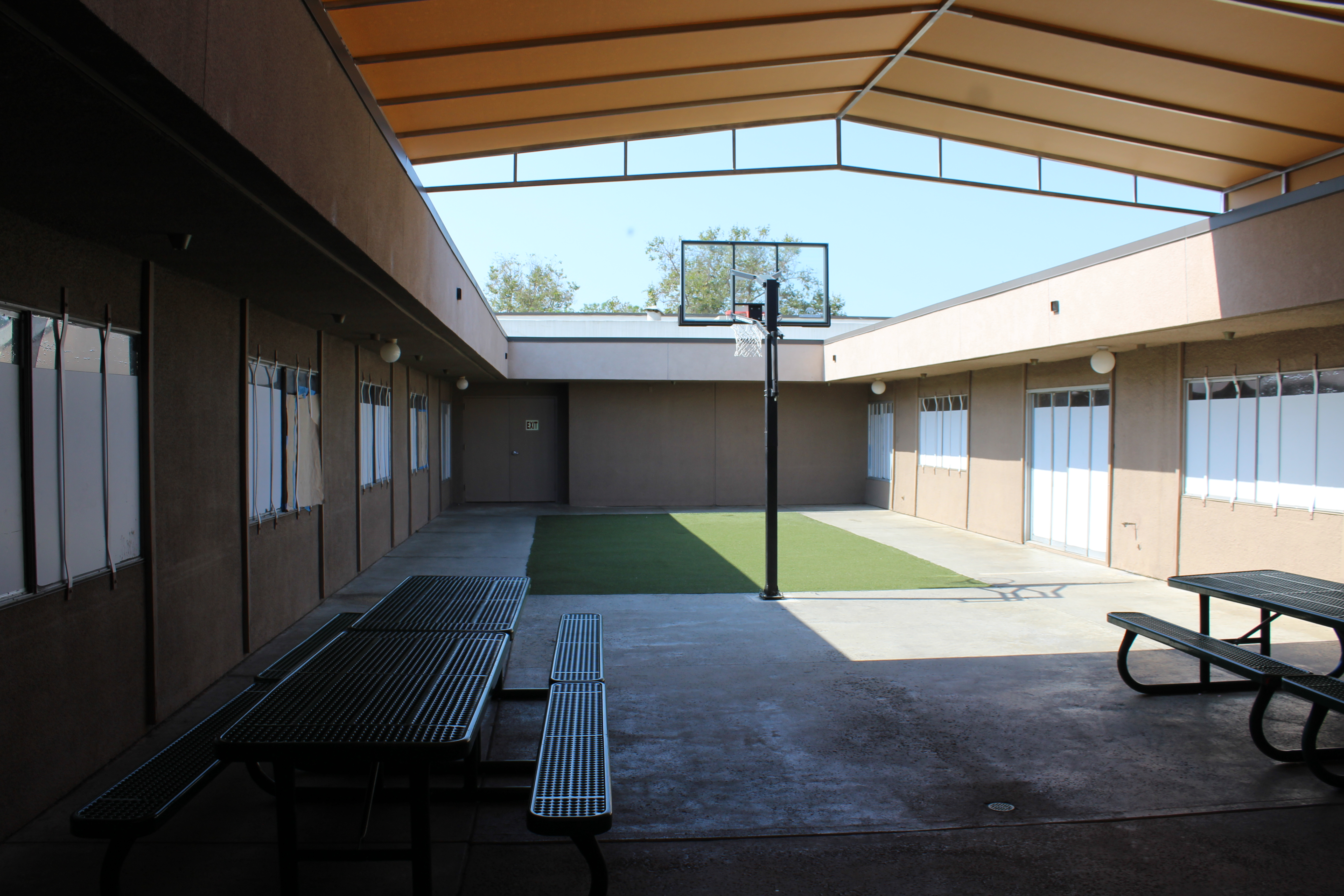A partially shaded, outdoor patio with 2 picnic tables and 1 basketball hoop. There is a rectangular area of astroturf facing the hoop. The patio is surrounded by walls with 9 windows that are covered by paper from the outside preventing view from inside or out.