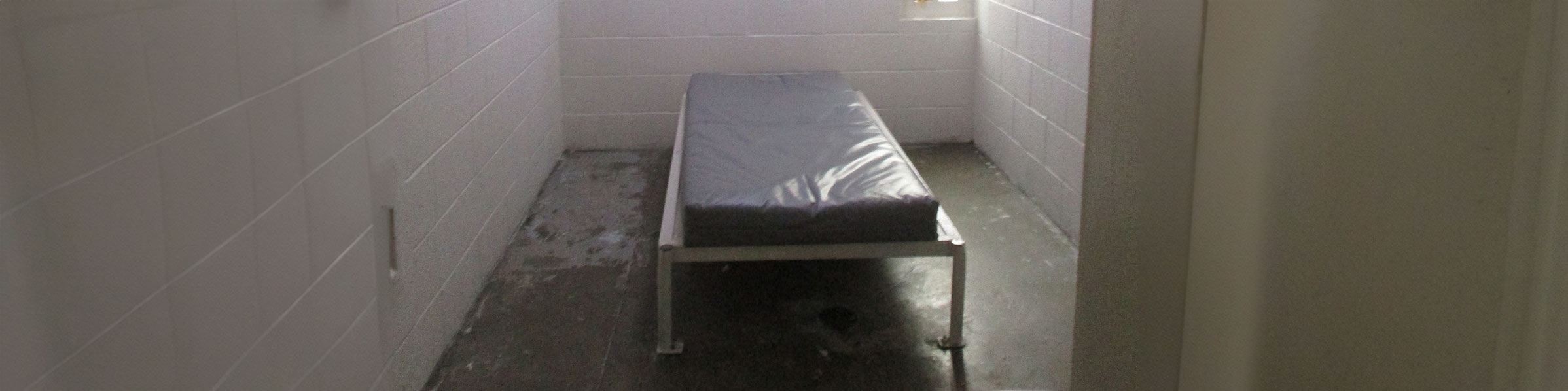 A cell at KCJC. The room has concrete floors and cinderblock walls, and is empty except for a bare mattress on a bed frame bolted to the floor and a metal toilet.