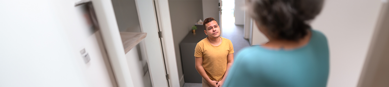 A latino man with dwarfism looking concerned as a latino woman neighbor is in front of his door.