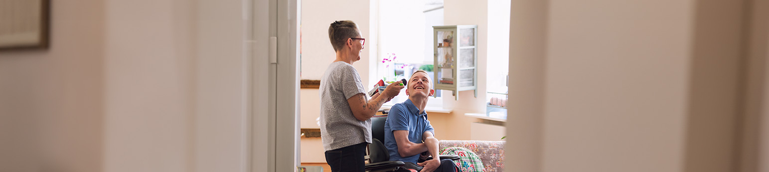 A white male with Cerebral Palsy in a wheelchair smiling together with a white female in their home.