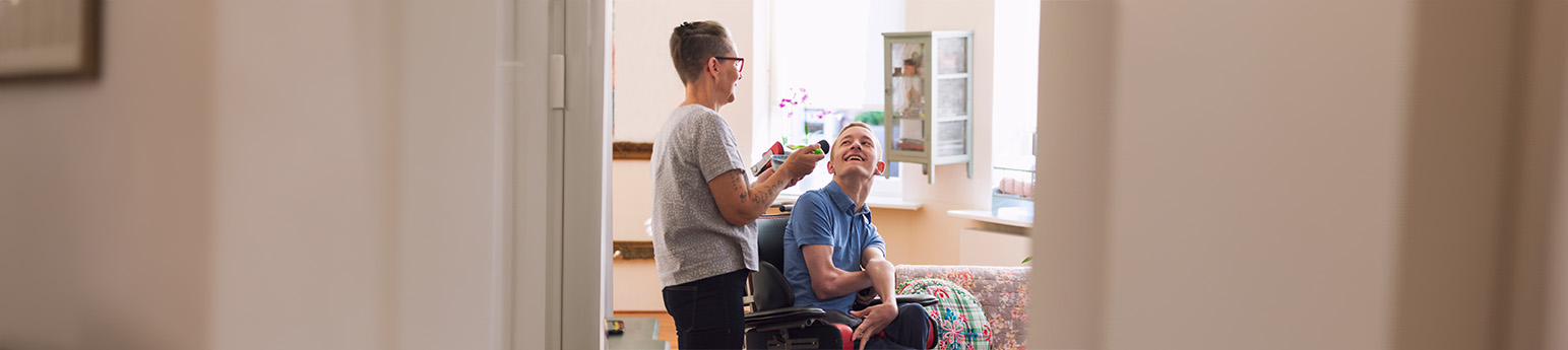 A white male with Cerebral Palsy in a wheelchair smiling together with a white female in their home.