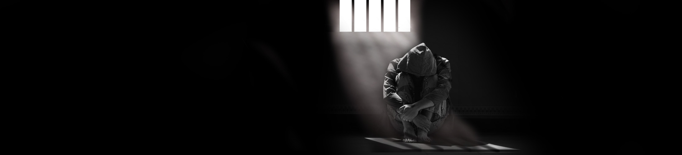 Light and shadow on surface of hopeless man in hoody sitting alone with hugging his knees on the floor in empty dark room in black and white style