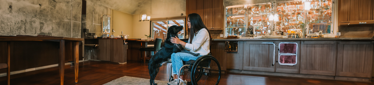 Woman of Asian ethnicity in her thirties with a physical disability, using a wheelchair pets her domestic dog in the kitchen of her home.
