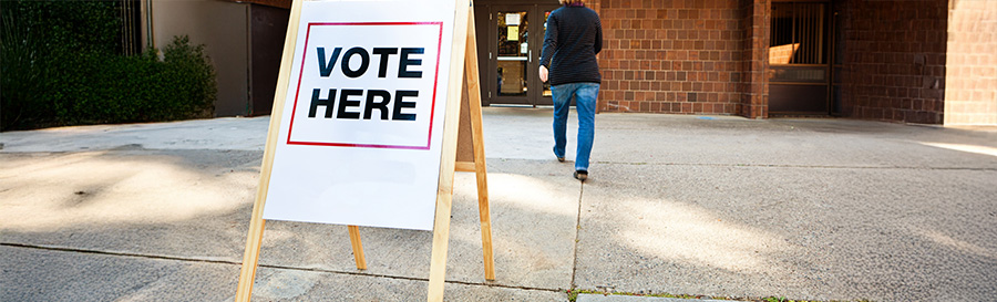 A brick building polling place with a stand that says, "Vote Here".