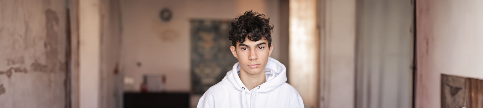 A teenage youth standing alone in a generally empty room looking depressed.