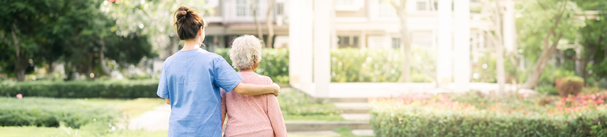 Caregiver walking with elderly woman outdoors in a beautiful, and relaxing, sun-lit garden.