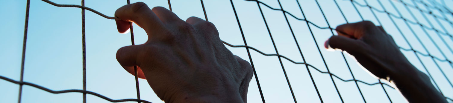 Close up photo of an older man's hands gripping a fence at a detention center.