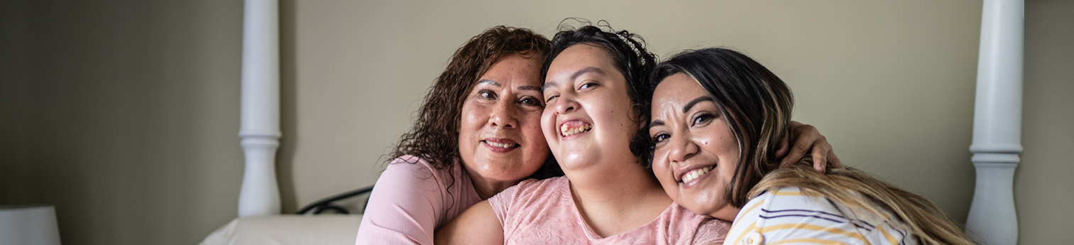 A Hispanic woman with a developmental disability smiling with her mother and her elder sister on either side of her.