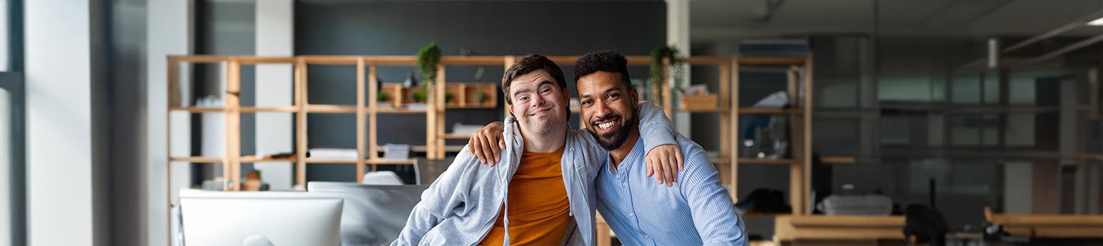 A white male with down syndrome and a black male smiling with a supportive side hug.