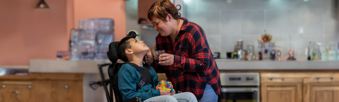 Hispanic IHHS woman caring for wheelchair-bound disabled boy in the kitchen, giving him a drink while smiling.