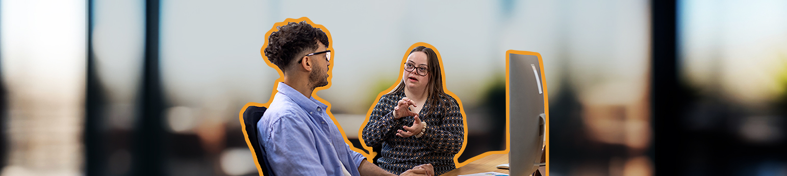Young adult white female with Down syndrome and her mid-adult latino colleague sitting at a desk in an office