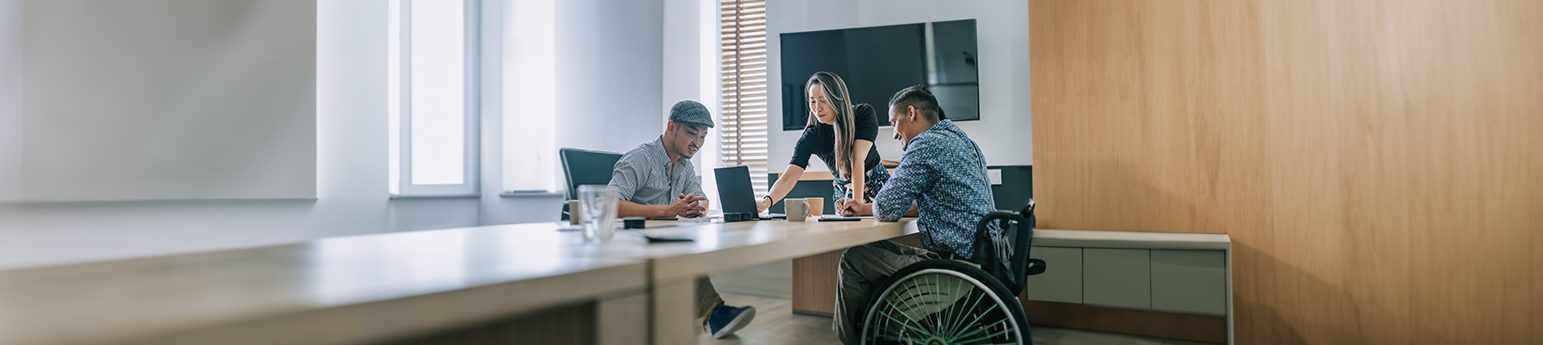 Three Asian individuals in a meeting room, one is a male in a wheelchair with darker skin, the other two, a man and woman with lighter skin, are helping him with job applications.