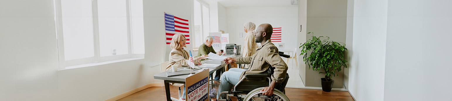 A Black man in a wheelchair asking voting questions to a White woman poll worker.
