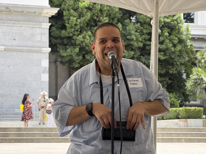 A blind speaker presenting at the event. He is a heavy set hispanic man who is blind and holding a braille assistant device.