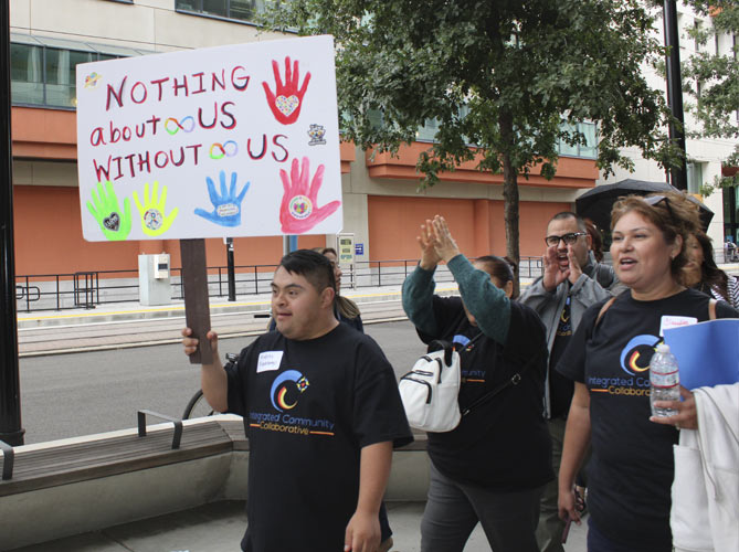 A crowd of people in a demonstration. They are crossing the street holding up a sign that saying, Nothing about us without us.