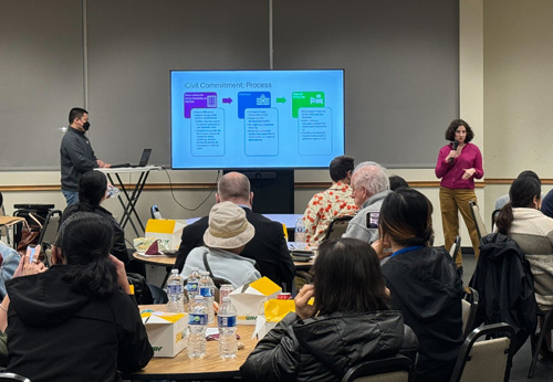 A young female DRC employee presenting at a community conversation. She is speaking to a room of people and behind her is a large monitor displaying her presentation.