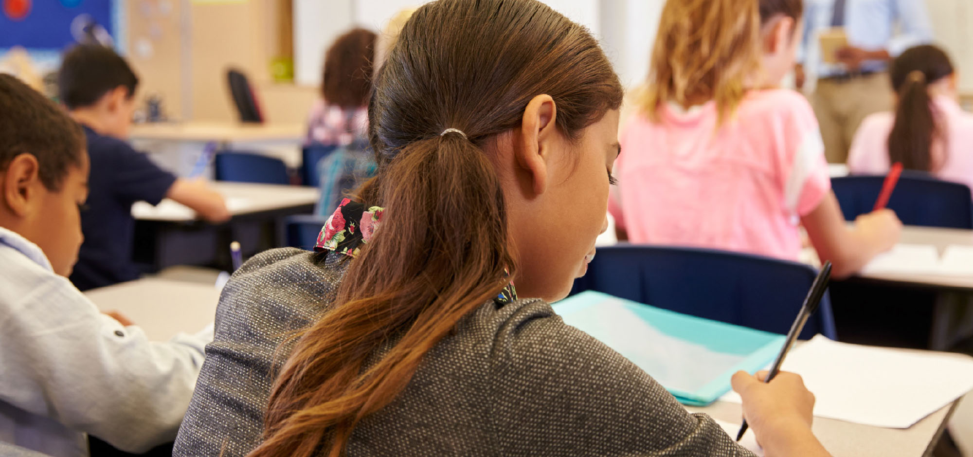 The back of a young girl sitting at her school desk with a full classroom of students.