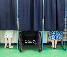 Image of three voting booths with cutains closed. The feet of two people can be seen below the curtain. One of the people voting is in a wheel chair.