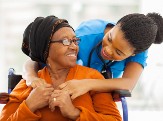 A nurse with here arms around an elderly woman sitting in a wheel chair. Their faces are looking toward each other and they are smiling.