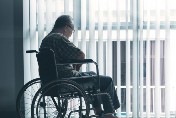 A lonely looking elderly man sitting in a wheel chair looking out a window.