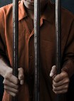 Image of a prisoner behind prison bars. He is gripping the bars with both hands. His face is hidden.