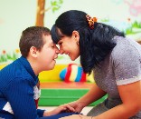 A female teacher leaning her forhead on the forhead of a young boy with a mental disability. They are in a classroom and are both smiling.