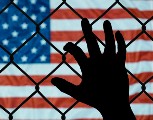 An image of a hand grabbing a chain linked fence. In the background is the American flag.