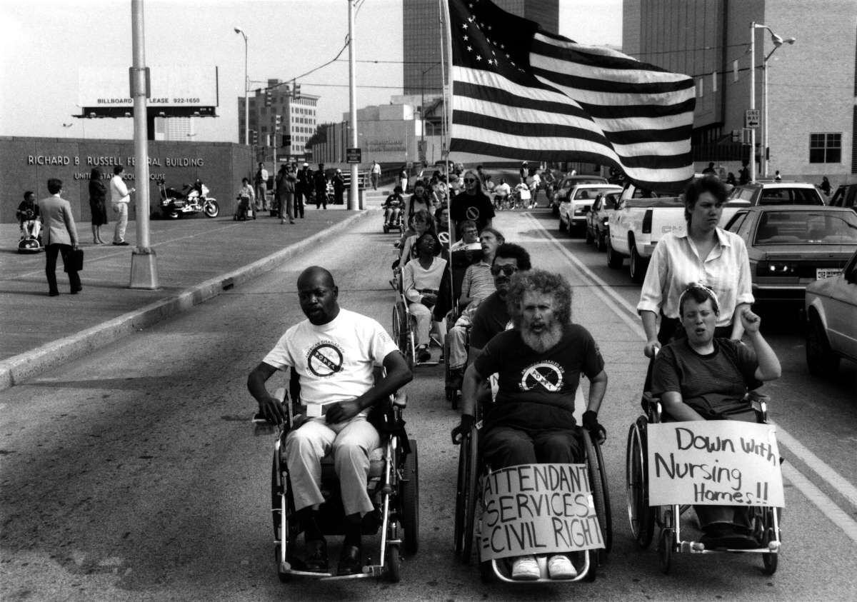 A black and white photo of a group of 3 disabled people in wheelchairs leading the march waving a United States Flag but the stars outline the disabled logo.
