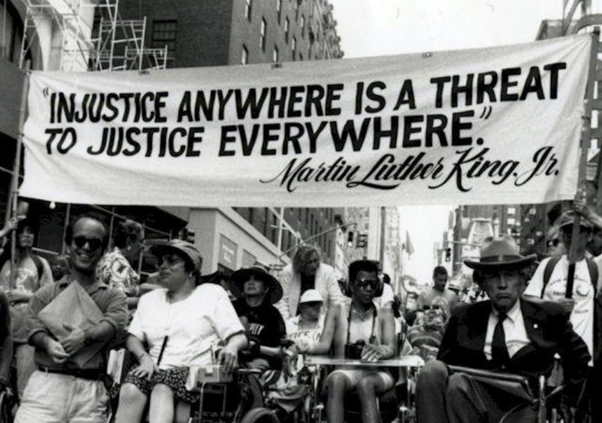 A black and white photo of a disabled marchers holding up a banner with Injustice anywhere is a threat to justice everywhere quoted by Martin Luther King Jr