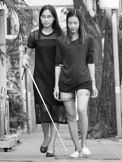 Photo of a blind woman with a walking cane walking on a sidewalk. She is being assisted by another woman.