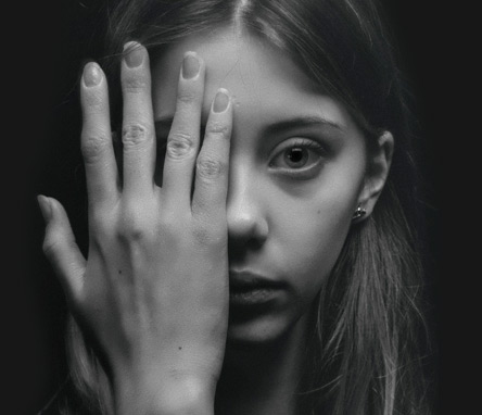 Close up image of a young woman's face who has a sad look. Her hand is covering half her face as she is directly looking at you.