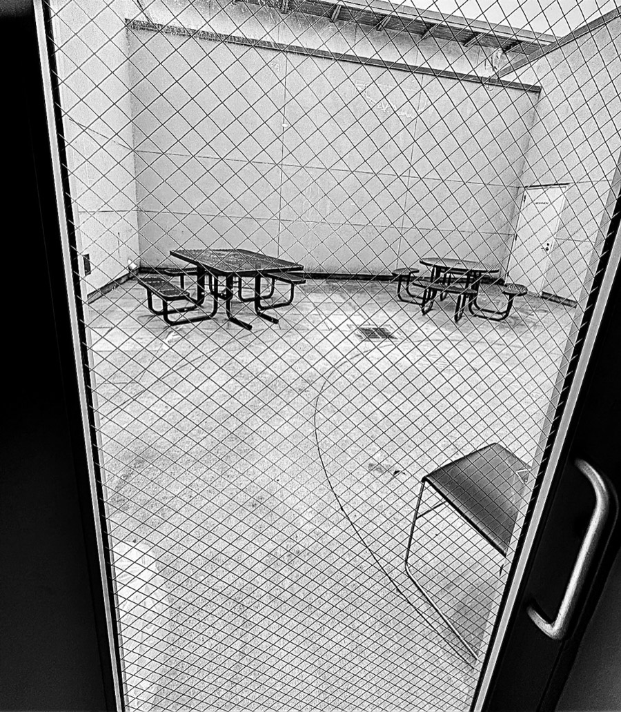 Looking through a metal door with safety glass there are metal picnic tables anchored down onto a concrete floor surrounded by beige painted cinderblock walls.