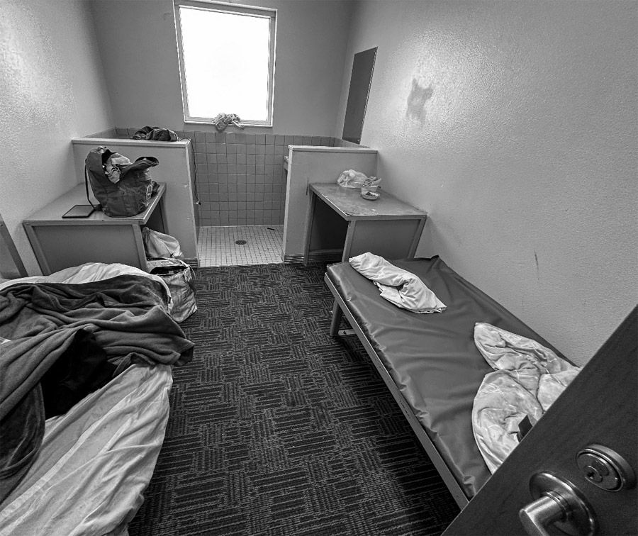 The view from the front of a double occupancy cell. In the foreground is the knob of the open door and two beds. The bed on the left is covered with rumpled blankets. The bed on the right has a plastic bed pad and some loose articles of clothing. A backpack and other personal items sit on metal desks behind the beds.