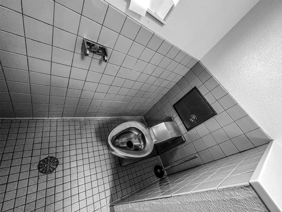 Close-up view of the bathroom section of a double occupancy cell. The floor is gray tile with a drain in the middle. Next to the carceral style metal toilet is a toilet paper holder and a standard issue plunger.