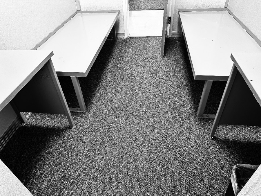 The view from the back of an unoccupied double-occupancy cell. In the foreground are two metal desks and a small plastic trash bin. In the background are two metal bed frames without bedding pads. The floor is a gray carpet fand the door is ajar.
