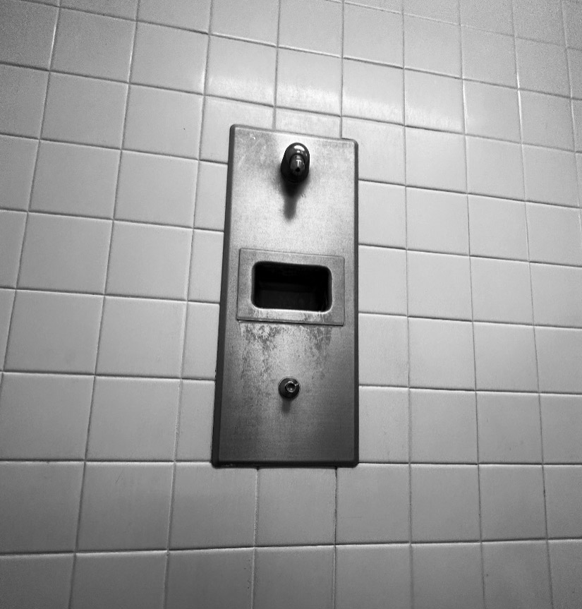 A close-up view of the push-button showerhead. Dull gray unpolished stainless-steel rectangle with a small shower spigot on top and a small button below.