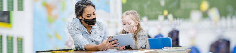 Image of a woman teaching a young girls who has a disability. They are both wearing COVID masks.