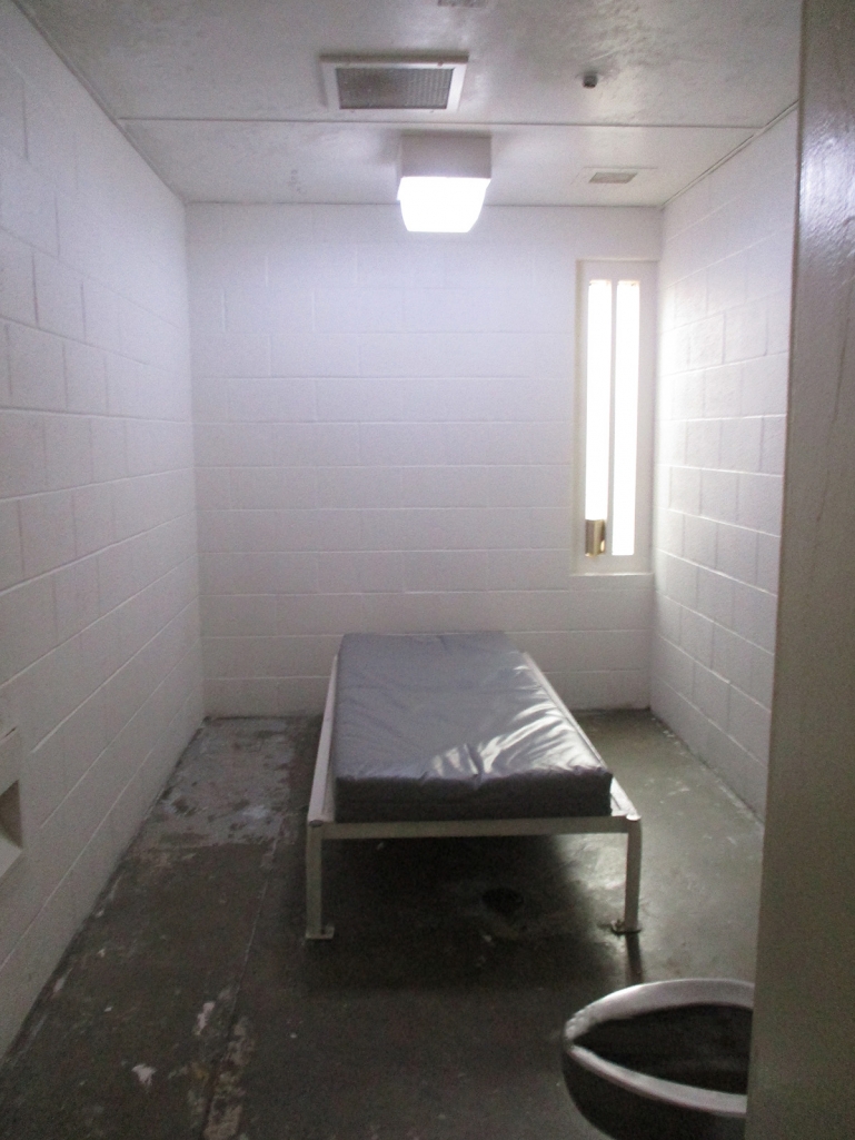 A cell at KCJC. The room has concrete floors and cinderblock walls, and is empty except for a bare mattress on a bedframe bolted to the floor and a metal toilet.